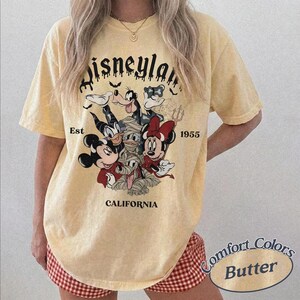 May include: A pale yellow short-sleeve t-shirt featuring a graphic of Disney characters in Halloween costumes. The text reads Disneyland Est 1955 California. The design includes Mickey Mouse, Minnie Mouse, Donald Duck, and Goofy in spooky attire.