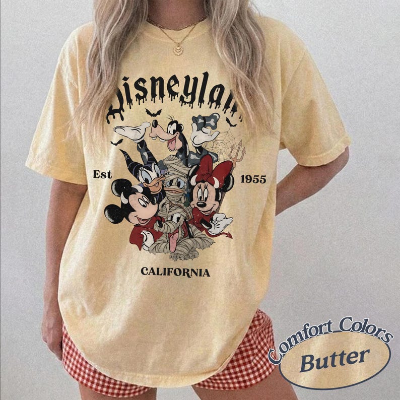 May include: A pale yellow short-sleeve t-shirt featuring a graphic of Disney characters in Halloween costumes. The text reads Disneyland Est 1955 California. The design includes Mickey Mouse, Minnie Mouse, Donald Duck, and Goofy in spooky attire.