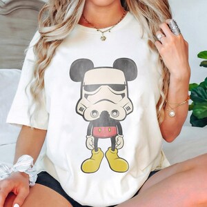 May include: Cream-colored t-shirt featuring a cartoon character mashup. The design combines Mickey Mouse's silhouette with a Stormtrooper helmet. The character wears a black body, red shorts, and yellow shoes. The shirt is a casual wear item.