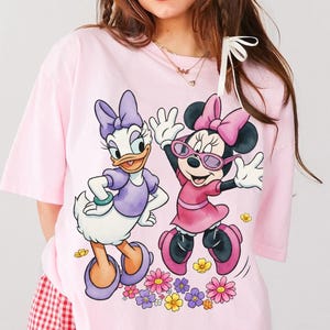 May include: A light pink t-shirt featuring Daisy Duck and Minnie Mouse. Daisy Duck is wearing a purple dress and Minnie Mouse is wearing a pink dress and sunglasses. The graphic includes colorful flowers at the bottom.