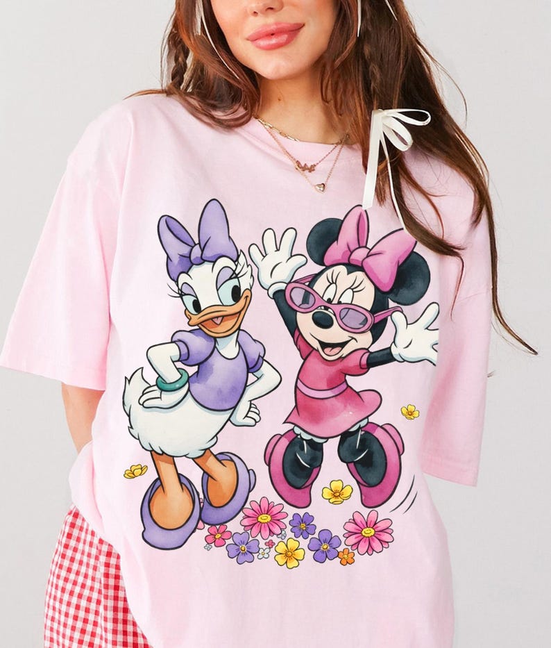 May include: A light pink t-shirt featuring Daisy Duck and Minnie Mouse. Daisy Duck is wearing a purple dress and Minnie Mouse is wearing a pink dress and sunglasses. The graphic includes colorful flowers at the bottom.