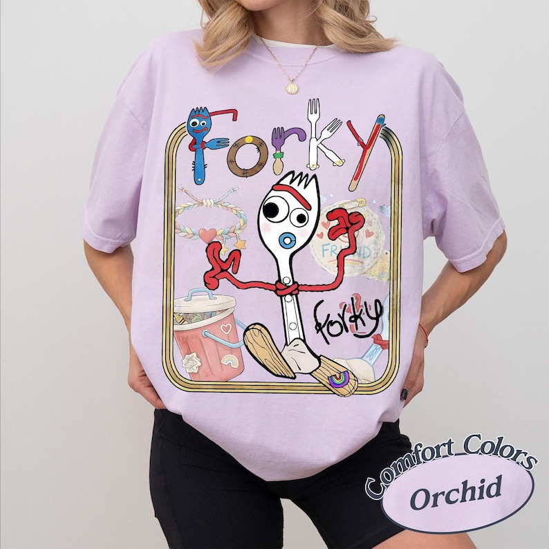 May include: A light purple short-sleeve t-shirt featuring a graphic print of the character Forky. The design includes the text Forky, a trash can, a bracelet, and various colourful illustrations enclosed within a rectangular gold-toned border.