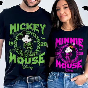 May include: Black t-shirts featuring Mickey and Minnie Mouse in Halloween skeleton designs. Mickey's shirt is green, and Minnie's is pink. Both shirts have the Disney logo and the year 1928.