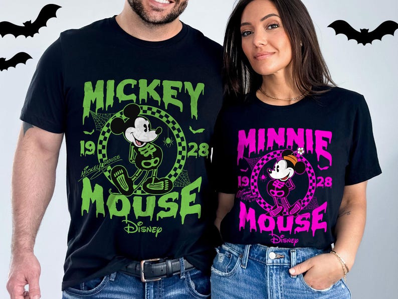 May include: Black t-shirts featuring Mickey and Minnie Mouse in Halloween skeleton designs. Mickey's shirt is green, and Minnie's is pink. Both shirts have the Disney logo and the year 1928.