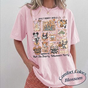 Pu&ograve; includere: T-shirt rosa chiaro con un design grafico di personaggi Disney in costume di Halloween. Il design include il testo "WALT DISNEY WORLD", "MICKEY'S Not-So-Scary Halloween Party" e "2026". La maglietta &egrave; etichettata "Comfort Colors Blossom".