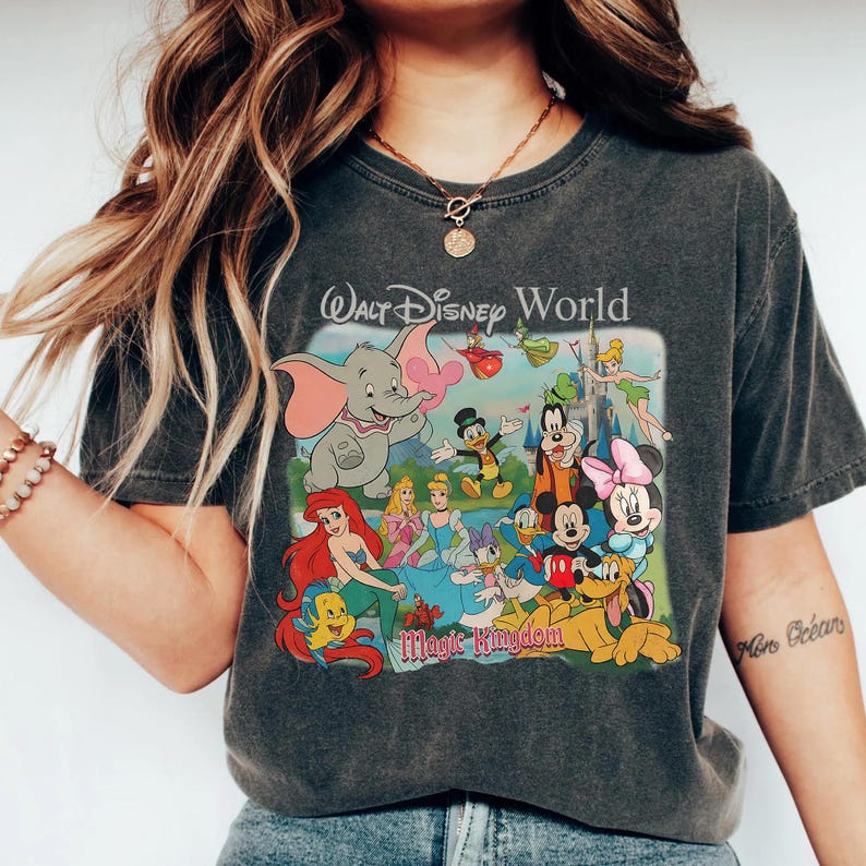 Pode incluir: T-shirt cinzenta escura com um gr&aacute;fico colorido de personagens da Disney, incluindo Dumbo, Mickey Mouse e Ariel. O texto "Walt Disney World Magic Kingdom" est&aacute; impresso acima e abaixo dos personagens. A camisa tem mangas curtas e gola redonda.