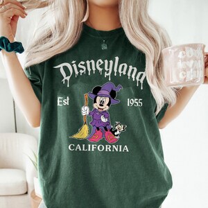 May include: Dark green t-shirt with a Disneyland graphic. The graphic features Minnie Mouse in a witch costume holding a broom, with a black cat. Text includes "Disneyland", "Est 1955", and "California".