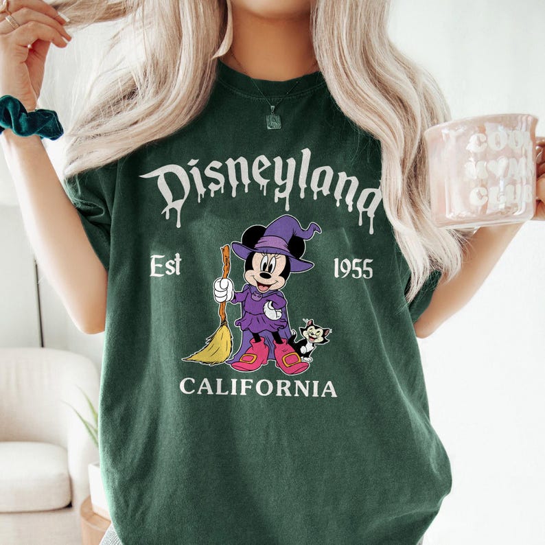 May include: Dark green t-shirt with a Disneyland graphic. The graphic features Minnie Mouse in a witch costume holding a broom, with a black cat. Text includes "Disneyland", "Est 1955", and "California".