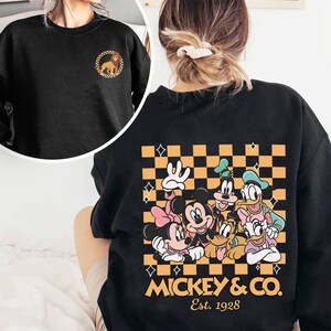 May include: Black sweatshirt with a checkered pattern and a graphic of Mickey Mouse and friends. The graphic includes Mickey Mouse, Minnie Mouse, Donald Duck, Goofy, Pluto, and Daisy Duck. The text "Mickey & Co. Est. 1928" is printed below the graphic.