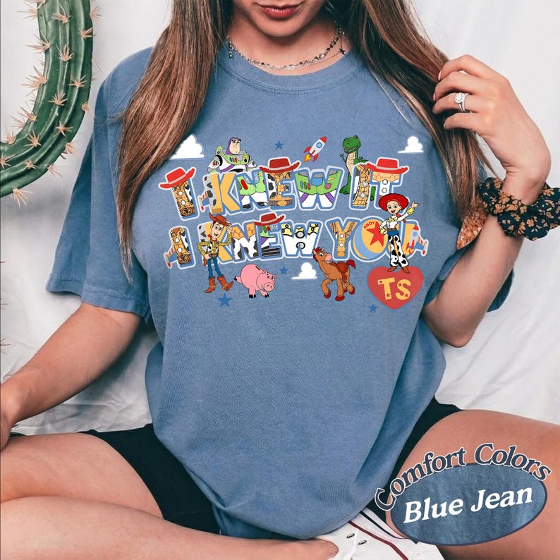 May include: A blue denim-colored t-shirt featuring a graphic print with the text I KNEW IT I KNEW YOU. The design includes cartoon characters like a cowboy, a space ranger, a cowgirl, a pig, and a horse, along with a rocket and a red heart with TS.
