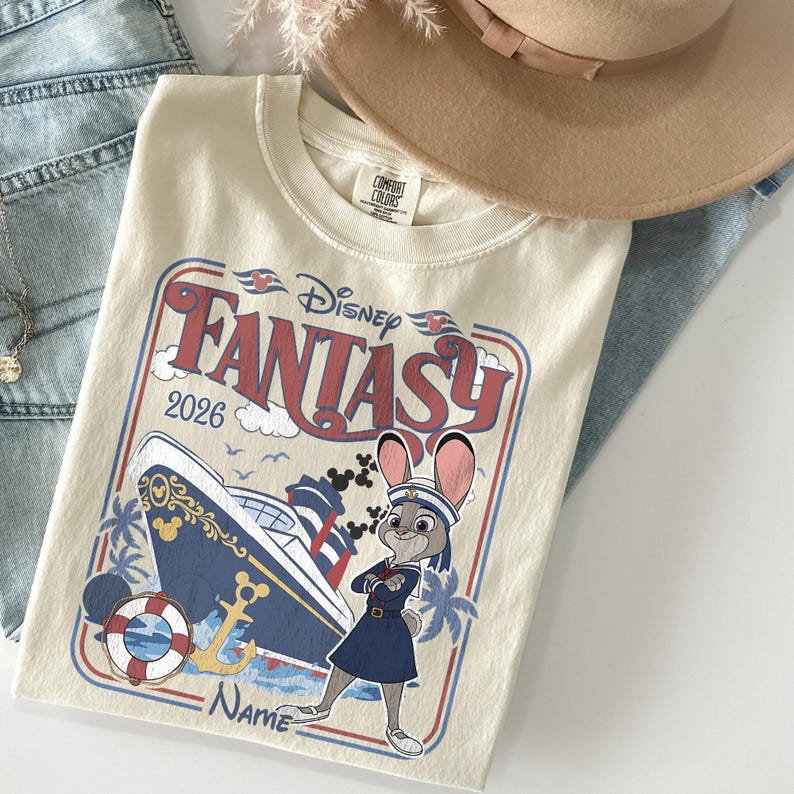 May include: Cream-colored t-shirt featuring a Disney Fantasy cruise ship design with the text "Disney Fantasy 2026." The graphic includes a cartoon character in a sailor outfit. The shirt is paired with blue jeans and a tan hat.