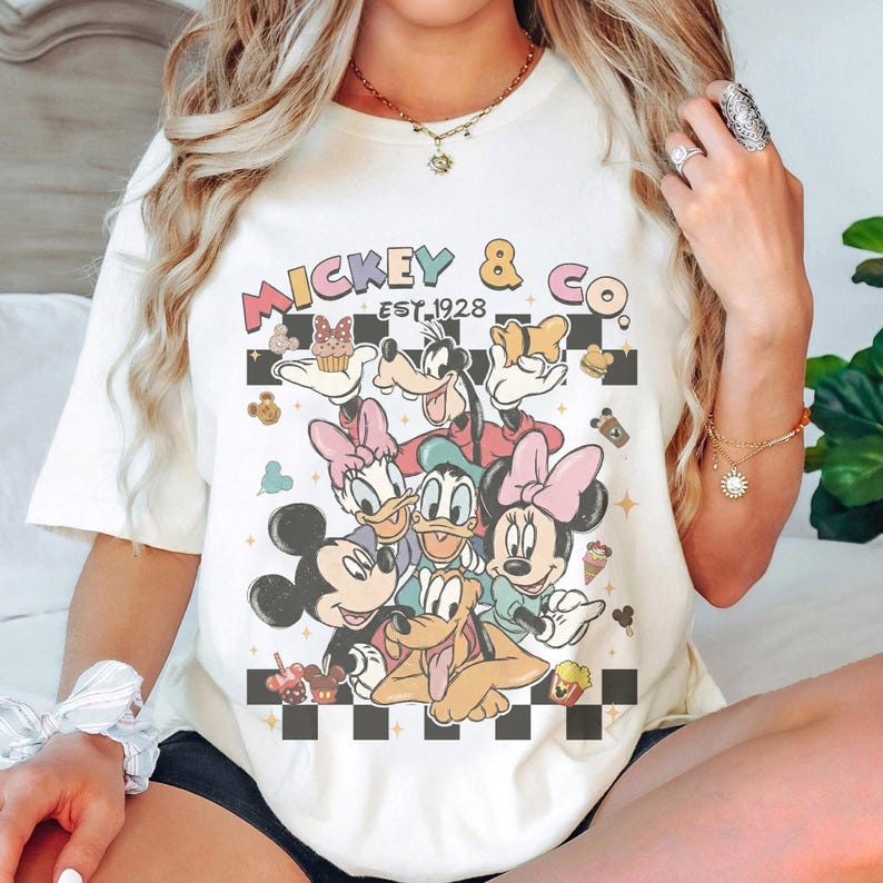 May include: Cream-colored t-shirt featuring a colorful graphic of Mickey Mouse, Minnie Mouse, Donald Duck, Daisy Duck, Goofy, and Pluto. The shirt has the text "Mickey & Co. EST. 1928" in a retro font, with a black and white checkered pattern.
