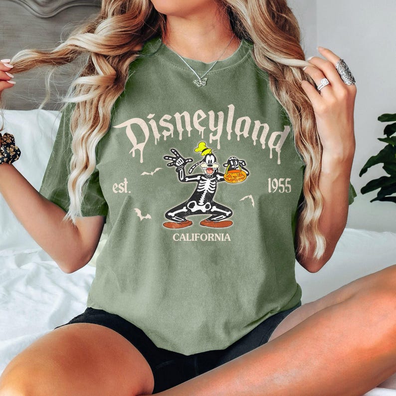 K&ouml;nnte beinhalten: Olivgr&uuml;nes T-Shirt mit einem Disneyland-Motiv. Das Motiv zeigt eine Skelett-Goofy-Figur mit einem K&uuml;rbis, den Text "Disneyland" und "est. 1955 CALIFORNIA".