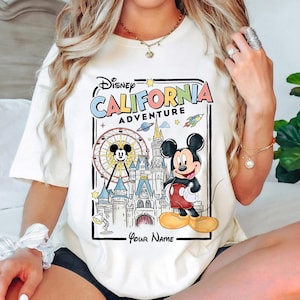 Puede incluir: Camiseta color crema con un gr&aacute;fico colorido de Mickey Mouse frente a un castillo y una noria, con el texto "Disney California Adventure". El dise&ntilde;o incluye estrellas, un cohete y las palabras "Your Name" en la parte inferior.