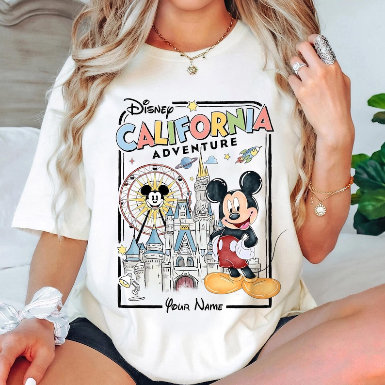 Puede incluir: Camiseta color crema con un gr&aacute;fico colorido de Mickey Mouse frente a un castillo y una noria, con el texto "Disney California Adventure". El dise&ntilde;o incluye estrellas, un cohete y las palabras "Your Name" en la parte inferior.