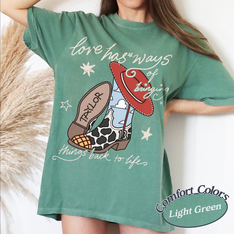May include: A light green t-shirt featuring a graphic of a cowboy boot with a cow print pattern, a blue sky with clouds, and the number 13. A red hat rests on the boot, and the text reads, love has ways of bringing things back to life.