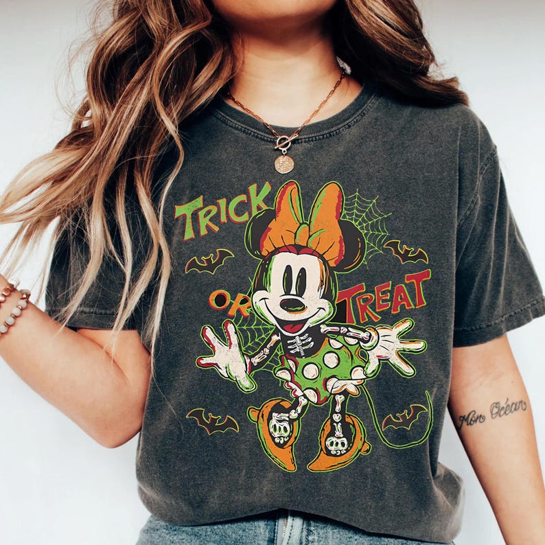 May include: Dark gray t-shirt featuring a Halloween-themed Minnie Mouse design. The graphic includes Minnie Mouse in a skeleton costume, a spider web, bats, and the words "Trick or Treat" in orange and green.