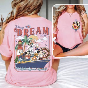 May include: Pink t-shirt featuring a Disney Dream cruise design. The back graphic includes Disney characters on a ship with a lighthouse and the text "DREAM 2026." The front has an anchor design with Mickey Mouse and the text "Name."