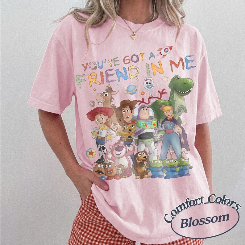 May include: A light pink t-shirt featuring a colorful graphic of various animated characters from the Toy Story franchise. The text "YOU'VE GOT A FRIEND IN ME" is printed above the characters in a multi-colored, playful font style.