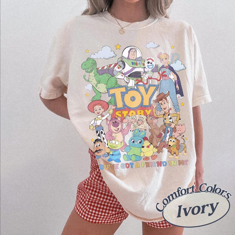 May include: An ivory short-sleeve t-shirt featuring a colorful graphic print of Toy Story characters including Woody, Buzz Lightyear, and Jessie. The text reads TOY STORY and YOU'VE GOT A FRIEND IN ME. The shirt is made by Comfort Colors.