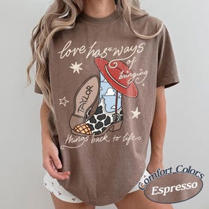 May include: A brown short-sleeve t-shirt featuring a graphic of a cowboy boot with a cow print pattern, a red hat, and the text "love has ways of bringing things back to life". The boot includes the name "TAYLOR" and the number "13".