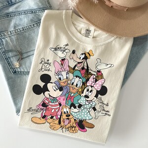 May include: Cream-colored t-shirt featuring a graphic of Disney characters including Mickey Mouse, Minnie Mouse, Donald Duck, Daisy Duck, Goofy, and Pluto. The shirt has the words "Mickey Mouse" and "Minnie Mouse" written on it. The shirt is folded on top of a pair of blue jeans.