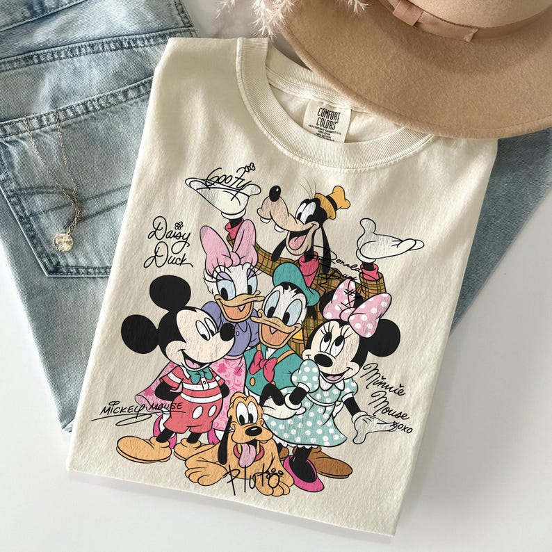 May include: Cream-colored t-shirt featuring a graphic of Disney characters including Mickey Mouse, Minnie Mouse, Donald Duck, Daisy Duck, Goofy, and Pluto. The shirt has the words "Mickey Mouse" and "Minnie Mouse" written on it. The shirt is folded on top of a pair of blue jeans.