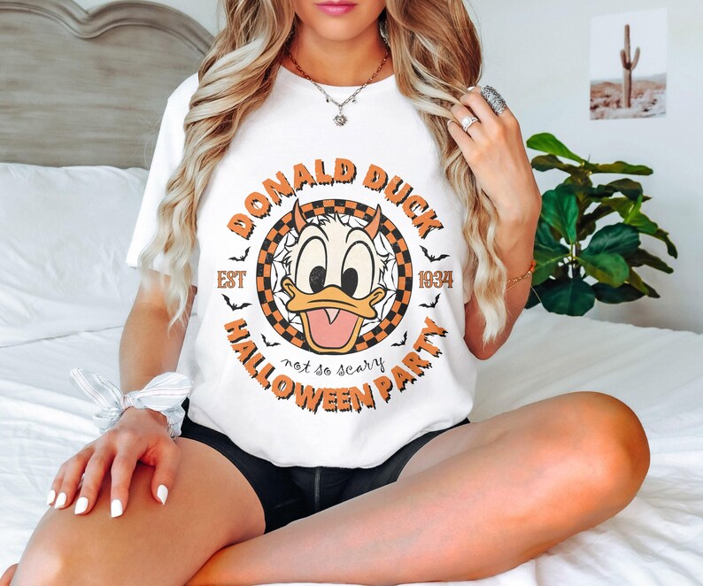 K&ouml;nnte beinhalten: Wei&szlig;es T-Shirt mit einem Aufdruck von Donald Duck und dem Text "Donald Duck Est 1934 Not so scary Halloween Party".