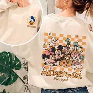 May include: A cream colored sweatshirt with a checkered pattern and a Mickey Mouse and friends graphic on the back. The graphic includes Mickey Mouse, Minnie Mouse, Donald Duck, Goofy, Pluto, and Daisy Duck. The text "Mickey & Co. Est. 1928" is printed below the graphic. The front of the sweatshirt has a smaller graphic of Donald Duck with the word "Brother" printed below it.