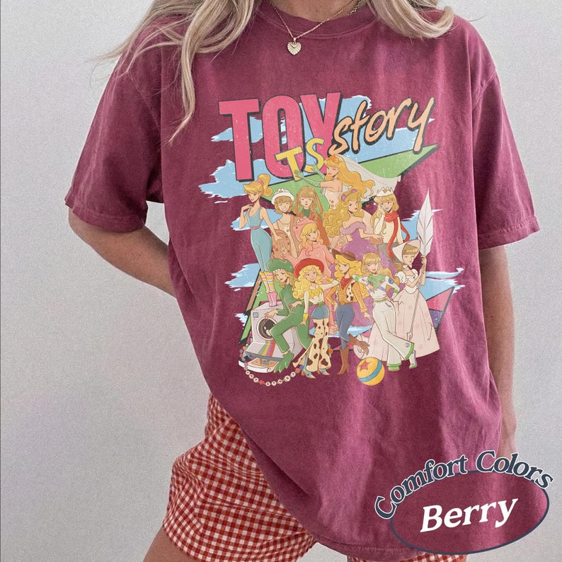 May include: A berry-colored short-sleeve t-shirt featuring a graphic print with the text "TOY story" in large pink and yellow letters. The design includes a collage of various animated characters in colorful outfits, set against a blue and green background.