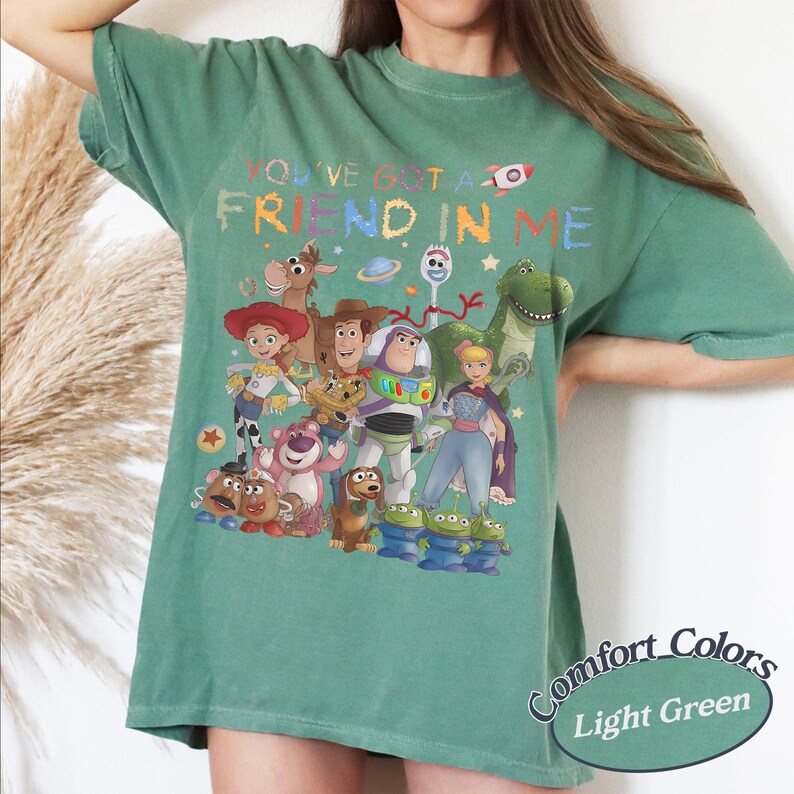 May include: A light green t-shirt featuring a colorful graphic print of various animated characters from the Toy Story franchise. The text "YOU'VE GOT A FRIEND IN ME" is printed above the characters in a multi-colored, playful font style.