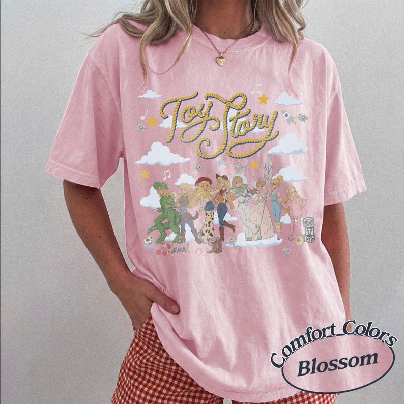 May include: A light pink short-sleeve t-shirt featuring the text Toy Story in yellow rope-style lettering. The graphic includes various animated characters, white clouds, stars, and musical notes. The shirt has a crew neckline and a relaxed fit.