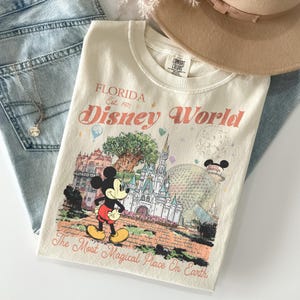 May include: Cream-colored t-shirt featuring a Disney World graphic with Mickey Mouse, a castle, and the text "Florida Est. 1971 Disney World The Most Magical Place On Earth." The shirt is folded on top of blue jeans.