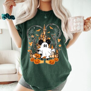 May include: Dark green t-shirt with a Halloween design. The graphic features a castle, Mickey Mouse ears, a ghost, pumpkins, bats, and fireworks. The shirt is a relaxed fit.