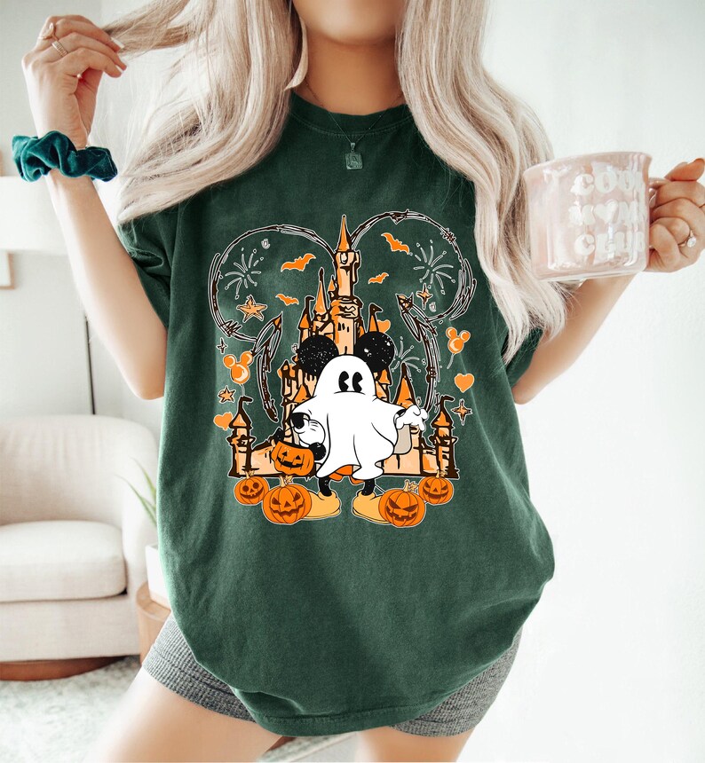 May include: Dark green t-shirt with a Halloween design. The graphic features a castle, Mickey Mouse ears, a ghost, pumpkins, bats, and fireworks. The shirt is a relaxed fit.