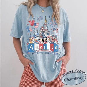 May include: A light blue short-sleeve t-shirt featuring a graphic of a castle, fireworks, and cartoon characters in red, white, and blue. The text reads America, 250 Years of Freedom, and 1776-2024. The shirt has a crew neckline and a relaxed fit.