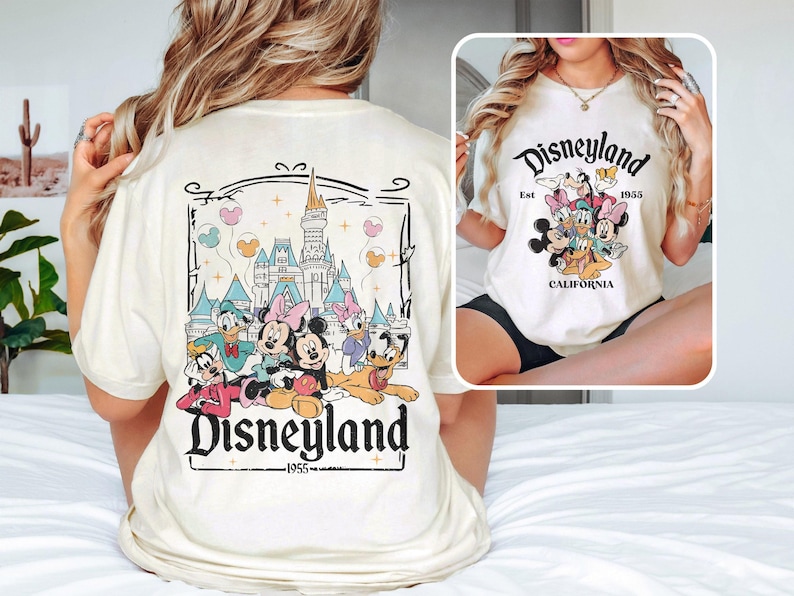 May include: White t-shirt with a graphic of Mickey Mouse and friends in front of Sleeping Beauty's castle. The text "Disneyland" is printed on the shirt with the year "1955" underneath.