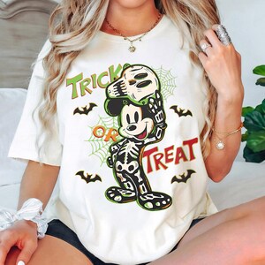 May include: Cream-coloured t-shirt with a Halloween-themed Mickey Mouse design. Mickey is dressed as a skeleton and holding a skull mask. The words "Trick or Treat" are printed in green and red, with bat and spiderweb graphics.