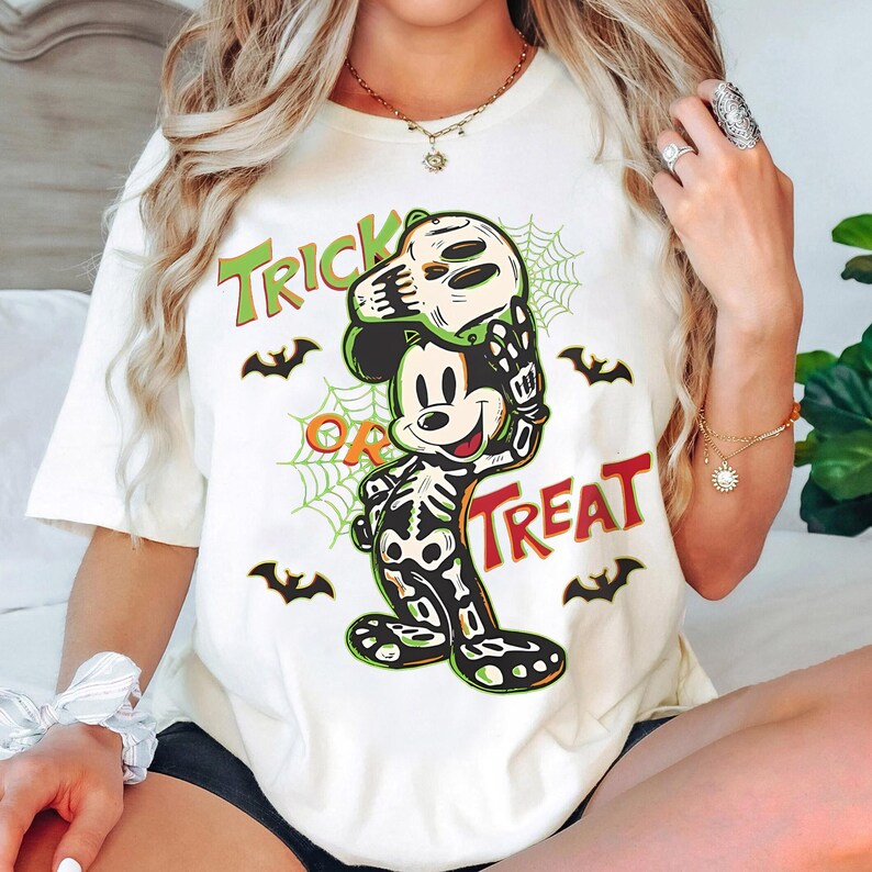 May include: Cream-coloured t-shirt with a Halloween-themed Mickey Mouse design. Mickey is dressed as a skeleton and holding a skull mask. The words "Trick or Treat" are printed in green and red, with bat and spiderweb graphics.
