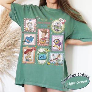 May include: A sage green t-shirt featuring a graphic print of various characters and items from the Toy Story franchise arranged in postage stamp-style frames, with the text "You've Got A Friend In Me" and "Hi!" printed in the center of the design.