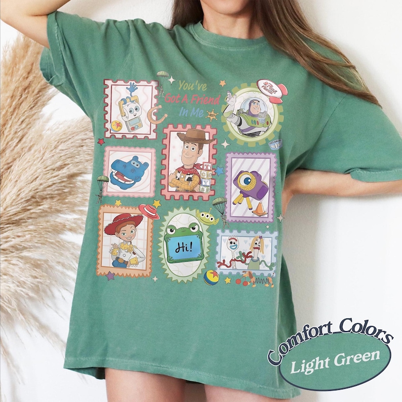 May include: A sage green t-shirt featuring a graphic print of various characters and items from the Toy Story franchise arranged in postage stamp-style frames, with the text "You've Got A Friend In Me" and "Hi!" printed in the center of the design.
