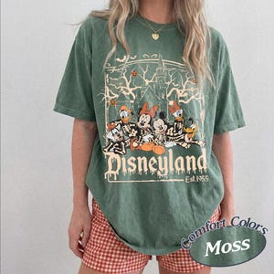 May include: Green t-shirt with a Disneyland graphic featuring Disney characters in skeleton costumes. The graphic includes a haunted house, pumpkins, and the text "Disneyland Est. 1955". The shirt is a Comfort Colors Moss color.