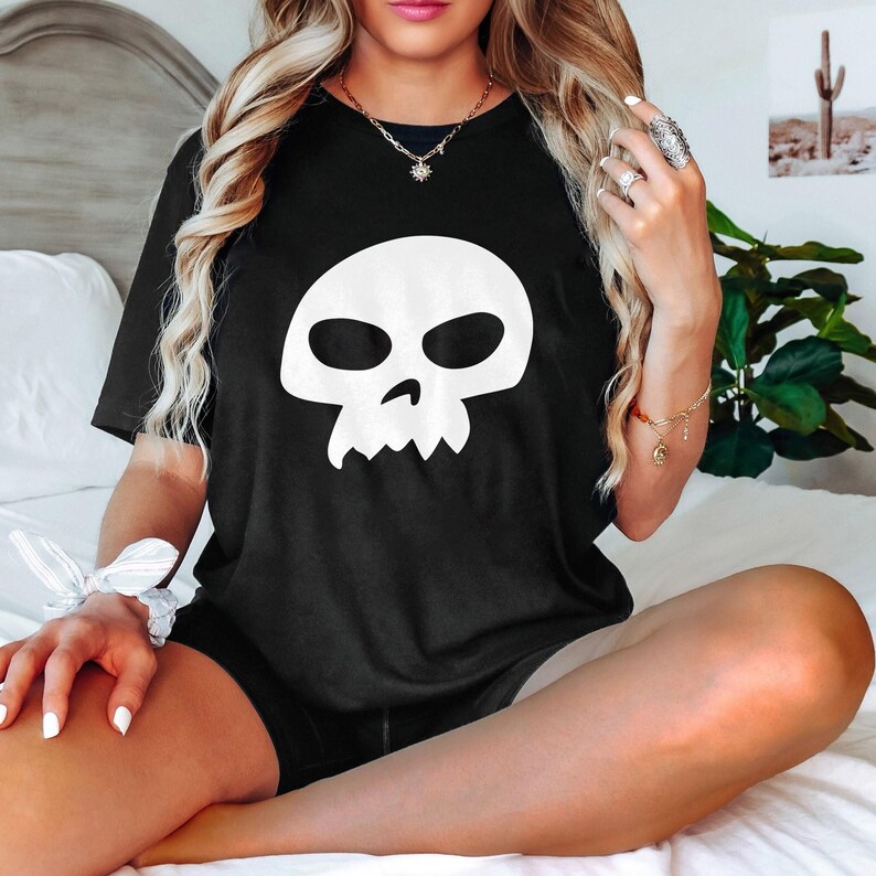 May include: Black t-shirt featuring a large white skull graphic. The skull design has large eye sockets and a jagged jawline. The shirt is short-sleeved and appears to be made of a soft material.