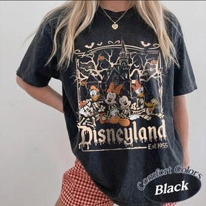 May include: Black t-shirt featuring a Halloween-themed Disneyland graphic. The design includes cartoon characters in skeleton costumes, a haunted house, bats, and pumpkins. The text "Disneyland Est. 1955" is also visible.