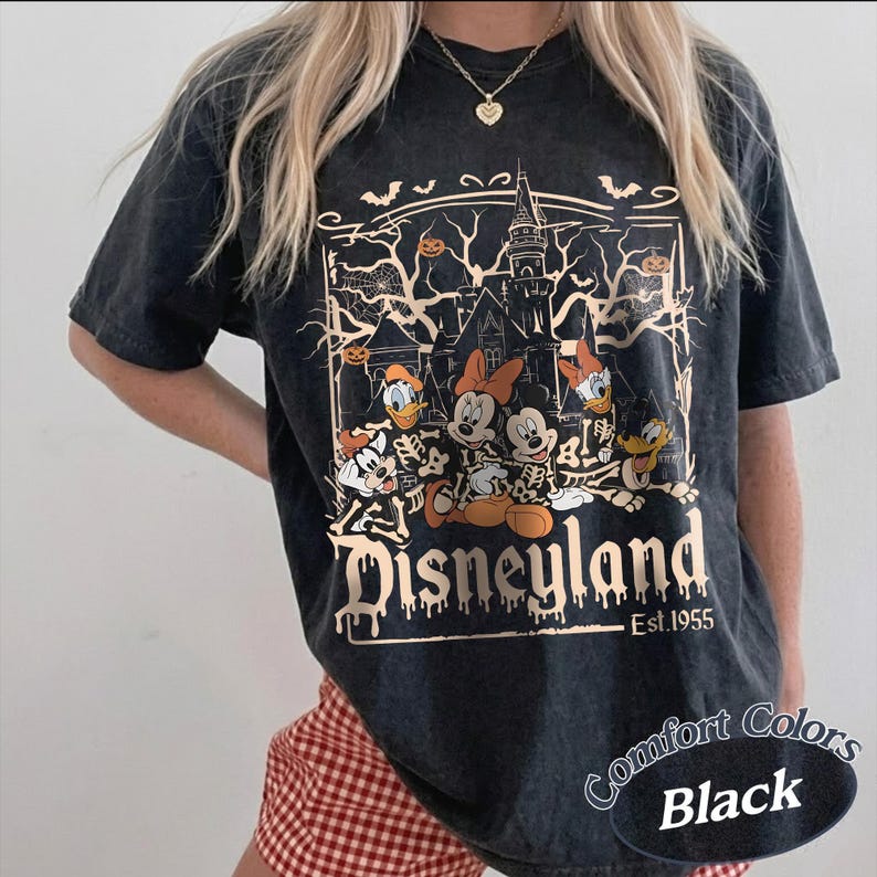 May include: Black t-shirt featuring a Halloween-themed Disneyland graphic. The design includes cartoon characters in skeleton costumes, a haunted house, bats, and pumpkins. The text "Disneyland Est. 1955" is also visible.