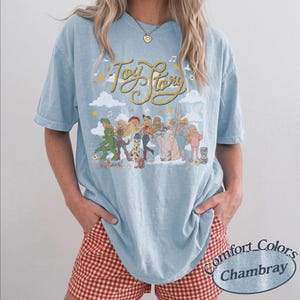 May include: A light blue short-sleeve t-shirt featuring a graphic print of various Toy Story characters arranged in a line. The text Toy Story is written in a yellow rope-style font above the characters, accompanied by clouds, stars, and musical notes.