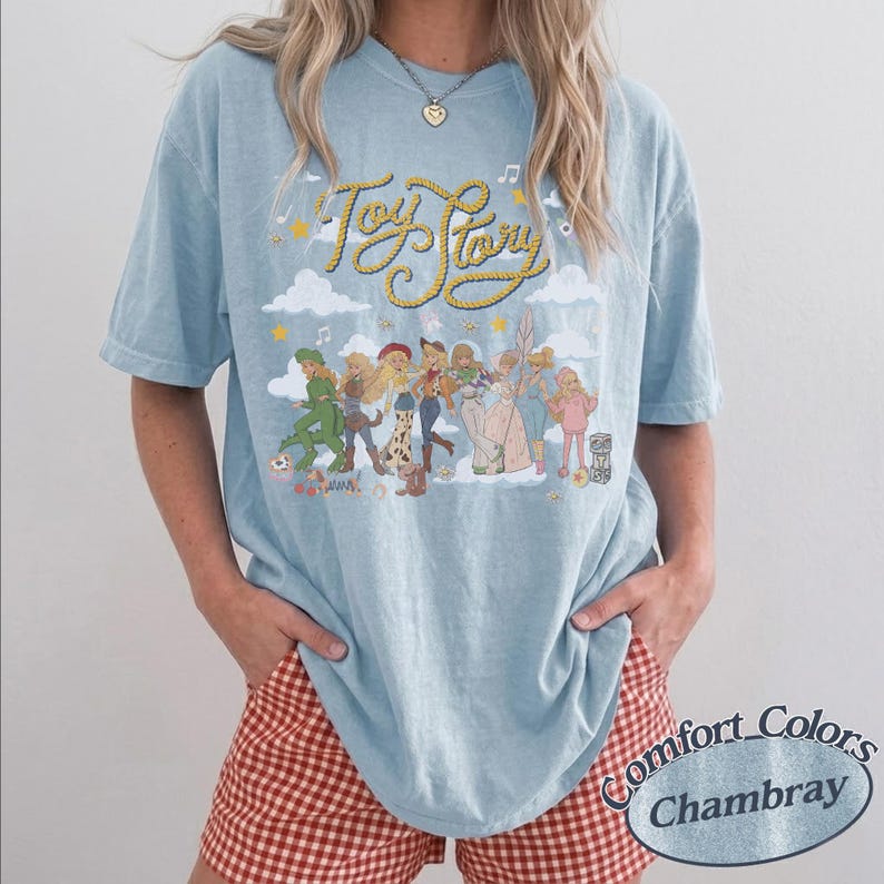 May include: A light blue short-sleeve t-shirt featuring a graphic print of various Toy Story characters arranged in a line. The text Toy Story is written in a yellow rope-style font above the characters, accompanied by clouds, stars, and musical notes.