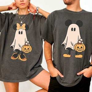 May include: Two black t-shirts with a ghost Mickey Mouse and Minnie Mouse design. The ghosts are holding jack-o'-lanterns. The shirts are a distressed charcoal color.