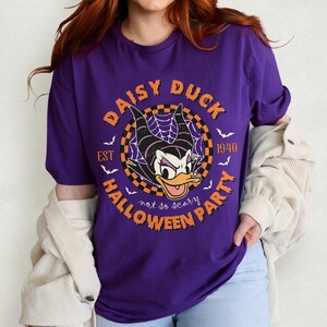 K&ouml;nnte beinhalten: Lilafarbenes T-Shirt mit einem Aufdruck von Daisy Duck als Hexe mit dem Text "Daisy Duck Halloween Party Est 1940 not so scary" in einem Kreis.