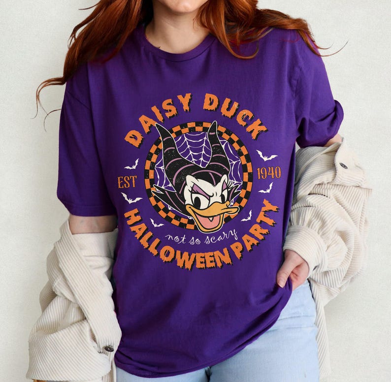K&ouml;nnte beinhalten: Lilafarbenes T-Shirt mit einem Aufdruck von Daisy Duck als Hexe mit dem Text "Daisy Duck Halloween Party Est 1940 not so scary" in einem Kreis.